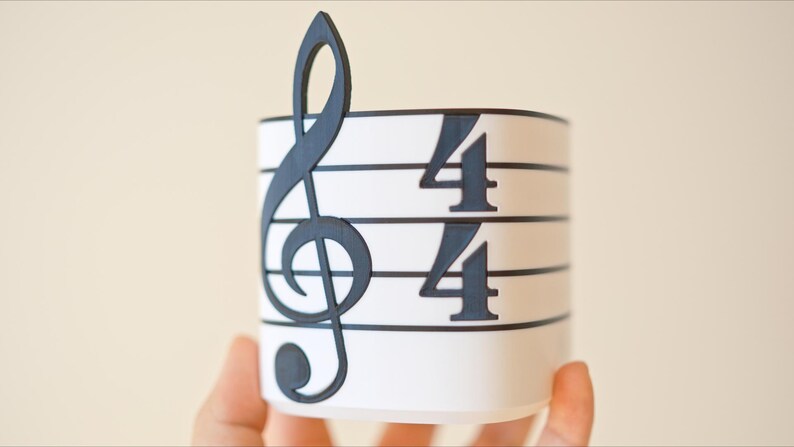 May include: A white cylindrical container with a black treble clef and 4/4 time signature design. The musical symbol is on the left side, and the time signature is on the right. The container is held by a person.
