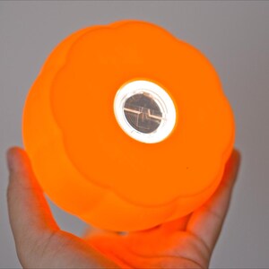 May include: Bright orange, spherical light with a central, circular, reflective element. The light has a textured surface and is held in a hand. The light is glowing, suggesting it is turned on. The overall shape is reminiscent of a pumpkin.
