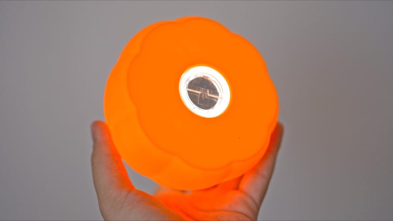 May include: Bright orange, spherical light with a central, circular, reflective element. The light has a textured surface and is held in a hand. The light is glowing, suggesting it is turned on. The overall shape is reminiscent of a pumpkin.