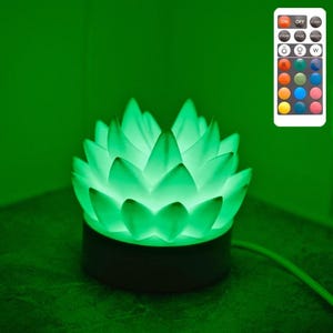 Op de afbeelding: Een groene LED-lamp in de vorm van een lotusbloem met een zwarte basis. De lamp is verlicht en werpt een groene gloed en een lotusbloemschaduw op de muur. Een afstandsbediening is zichtbaar.