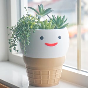 Op de afbeelding: Een witte en bruine keramische pot in de vorm van een ijshoorn met een glimlachend gezicht. De pot is gevuld met groene vetplanten.