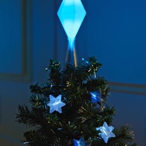 May include: A small Christmas tree with a blue diamond-shaped tree topper and glowing star-shaped lights. The tree is dark green and has a festive appearance, perfect for holiday decorations. The lights and topper emit a soft blue glow.