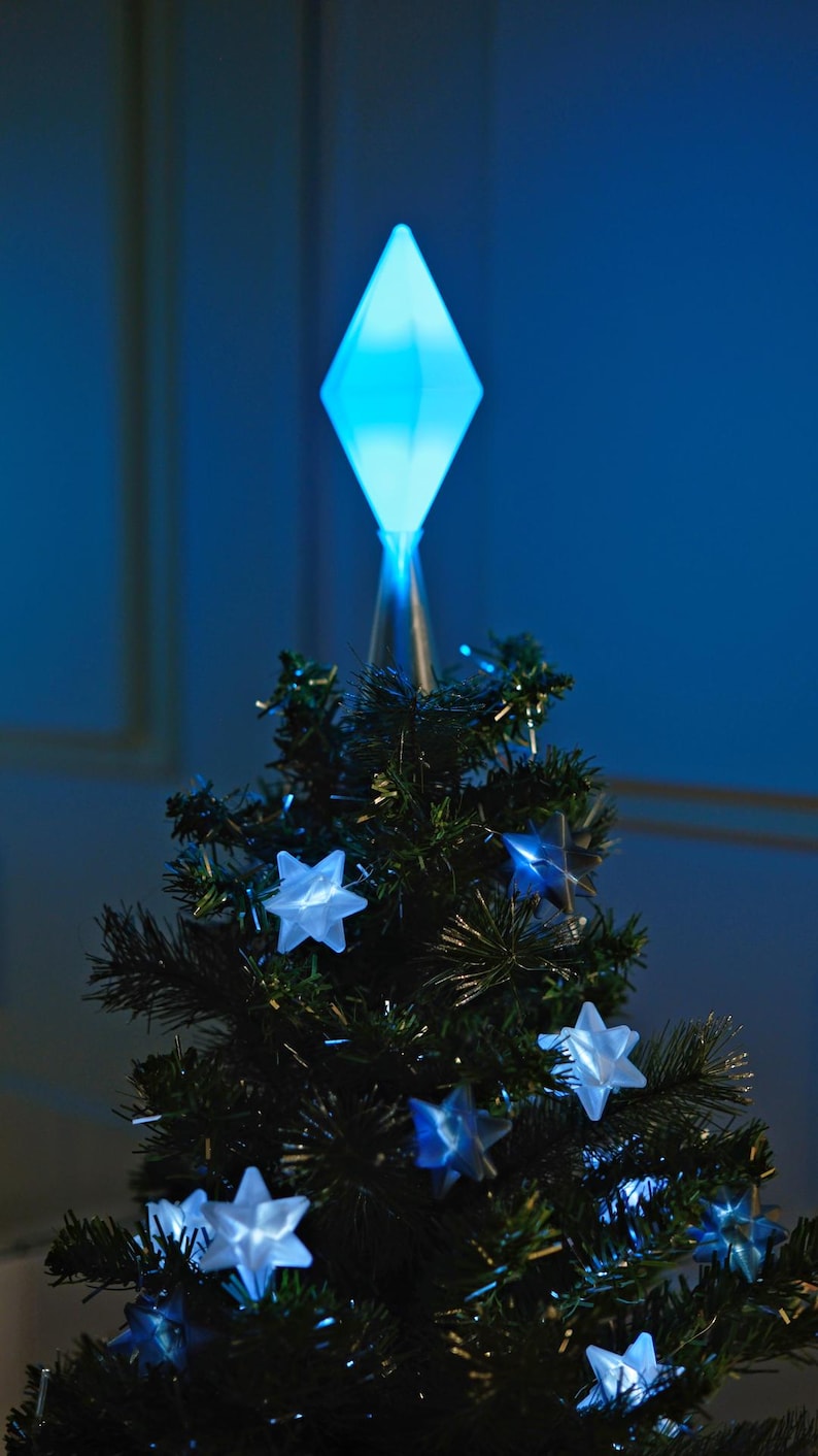 May include: A small Christmas tree with a blue diamond-shaped tree topper and glowing star-shaped lights. The tree is dark green and has a festive appearance, perfect for holiday decorations. The lights and topper emit a soft blue glow.