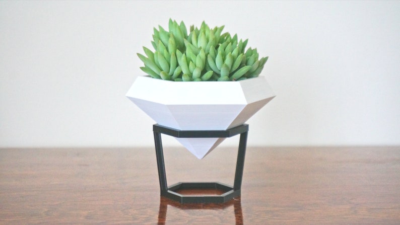 May include: A white geometric diamond-shaped planter with a black metal stand, filled with green succulent plants.