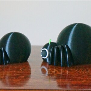 May include: Two black 3D printed spiders of different sizes. The larger spider is on the right and the smaller spider is on the left. Both spiders have green antennae and white eyes.