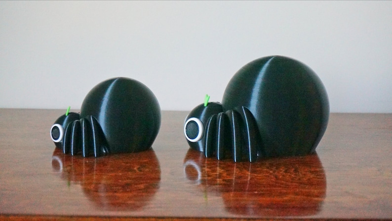 May include: Two black 3D printed spiders of different sizes. The larger spider is on the right and the smaller spider is on the left. Both spiders have green antennae and white eyes.