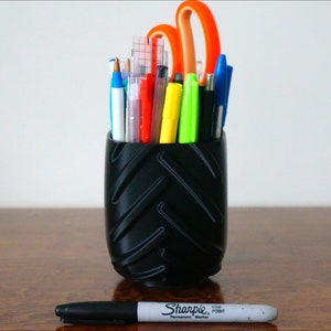 May include: A black tire-shaped pencil holder filled with various pens, pencils, and a pair of orange scissors. A black Sharpie permanent marker is lying on a wooden surface.