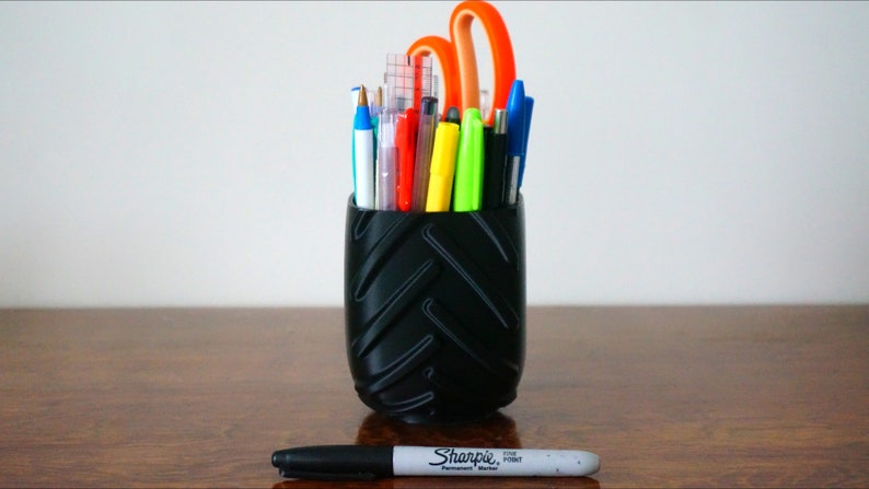 May include: A black tire-shaped pencil holder filled with various pens, pencils, and a pair of orange scissors. A black Sharpie permanent marker is lying on a wooden surface.