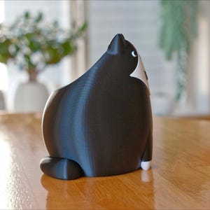 May include: A black and white cat figurine, viewed from the back, sits on a wooden table. The cat has a rounded body, with white markings on its chest and face. The figurine is a decorative item, possibly for a home or office.