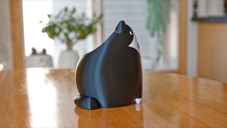 May include: A black and white cat figurine, viewed from the back, sits on a wooden table. The cat has a rounded body, with white markings on its chest and face. The figurine is a decorative item, possibly for a home or office.