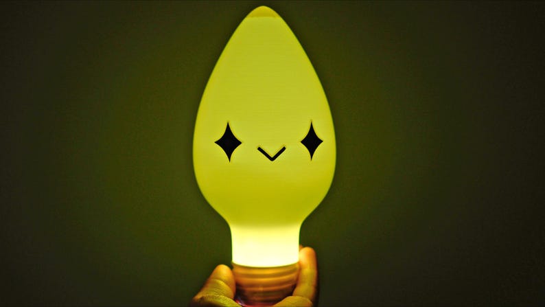 May include: A cheerful, yellow lightbulb with a cartoon face. The bulb has a teardrop shape and a standard base. The face features two black diamond-shaped eyes and a small, curved mouth. The light is bright.