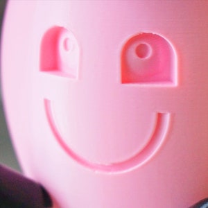 May include: A pink plastic object with a smiling face. The face has two eyes and a wide mouth.