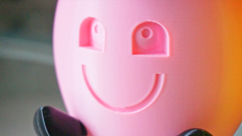 May include: A pink plastic object with a smiling face. The face has two eyes and a wide mouth.