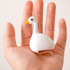 May include: A small, white goose figurine with an orange beak and black eyes. The figurine is held in an open hand against a plain background. The goose has a curved neck and a rounded body, with a small, curved tail.