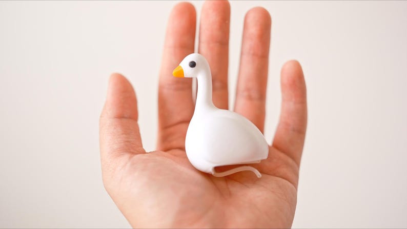 May include: A small, white goose figurine with an orange beak and black eyes. The figurine is held in an open hand against a plain background. The goose has a curved neck and a rounded body, with a small, curved tail.