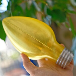 May include: A yellow decorative light bulb with a silver base. The bulb is held in front of a background of green leaves. The bulb is a unique decorative item.