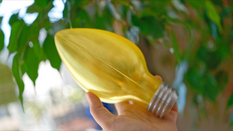 May include: A yellow decorative light bulb with a silver base. The bulb is held in front of a background of green leaves. The bulb is a unique decorative item.