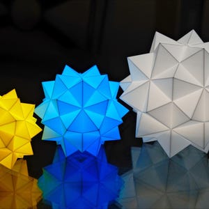 Pode incluir: Tr&ecirc;s estrelas de papel geom&eacute;tricas em tamanhos diferentes. A maior estrela &eacute; branca, a estrela do meio &eacute; azul e a menor estrela &eacute; amarela.