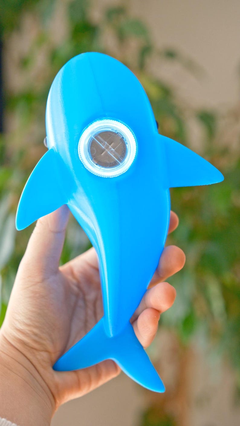 May include: A bright blue dolphin-shaped toy with a circular, clear window in the center. The toy has fins and a tail, and is held in a person's hand. The toy is made of a smooth, shiny material.