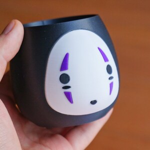 May include: A black and white ceramic cup featuring a No-Face design from the animated film Spirited Away. The cup has a rounded shape with a white face and purple details. The cup is held in a hand.