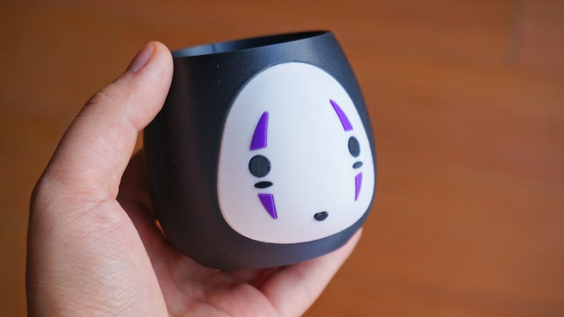 May include: A black and white ceramic cup featuring a No-Face design from the animated film Spirited Away. The cup has a rounded shape with a white face and purple details. The cup is held in a hand.