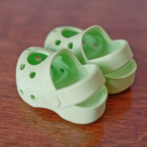 May include: Two light green plastic sandals with a hole pattern. The sandals have a strap that goes around the ankle.