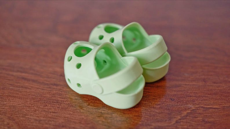 May include: Two light green plastic sandals with a hole pattern. The sandals have a strap that goes around the ankle.