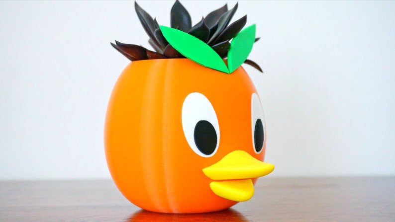 May include: Orange ceramic planter shaped like a duck with a green succulent plant inside.