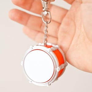 May include: A miniature drum keychain with a silver chain and clasp. The drum has a white face, a bright orange body, and silver trim. The keychain is held up against a plain background.