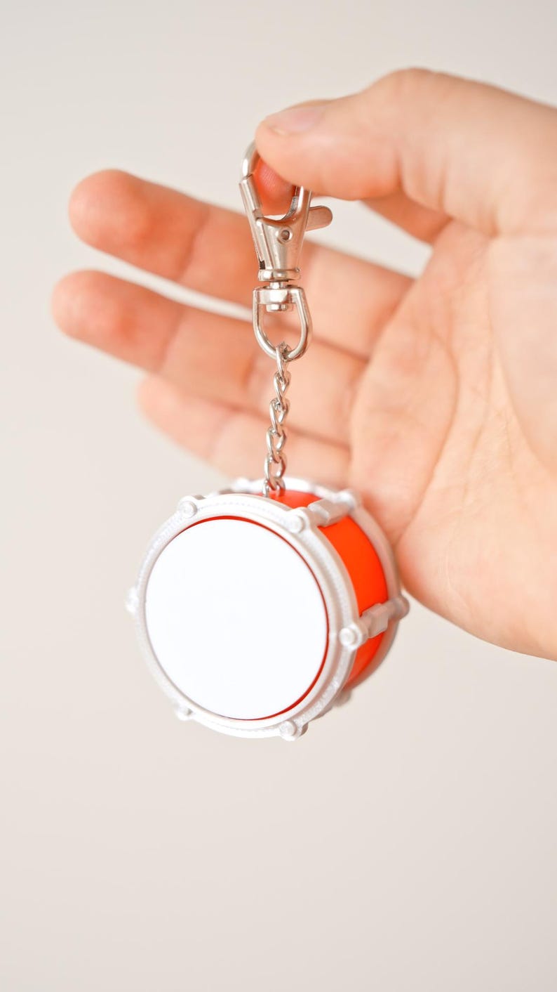May include: A miniature drum keychain with a silver chain and clasp. The drum has a white face, a bright orange body, and silver trim. The keychain is held up against a plain background.