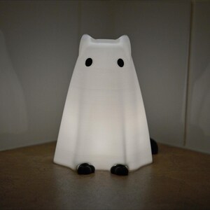 May include: A white ghost-shaped nightlight with black eyes. The light is on and glowing.