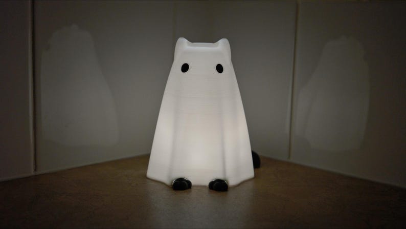 May include: A white ghost-shaped nightlight with black eyes. The light is on and glowing.