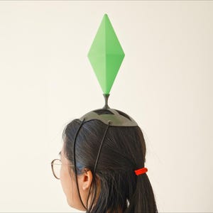 May include: A person wearing a headpiece with a green diamond-shaped object on top, resembling the plumbob from The Sims video game. The headpiece is dark gray, and the person is wearing glasses and has dark hair.