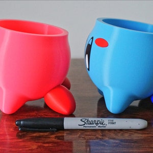 May include: Two 3D printed planters shaped like a pink and blue character with three legs. The character has a large round head and a small body. The planters are on a wooden surface.