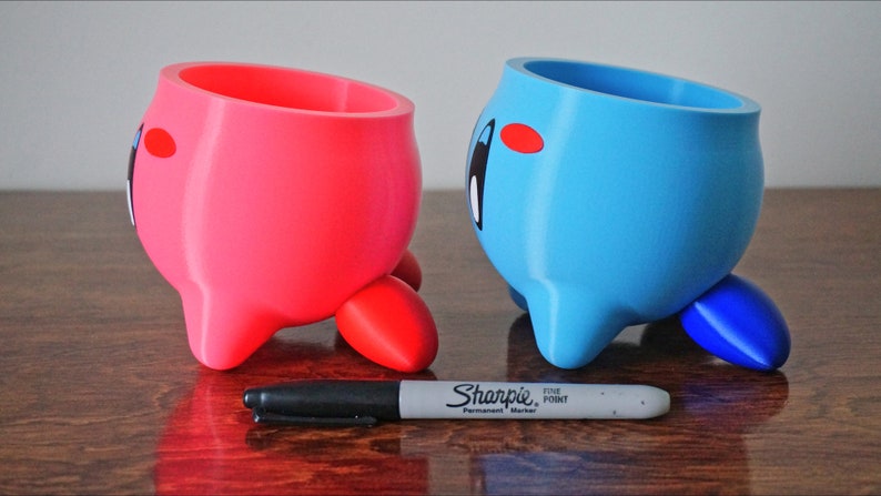 May include: Two 3D printed planters shaped like a pink and blue character with three legs. The character has a large round head and a small body. The planters are on a wooden surface.