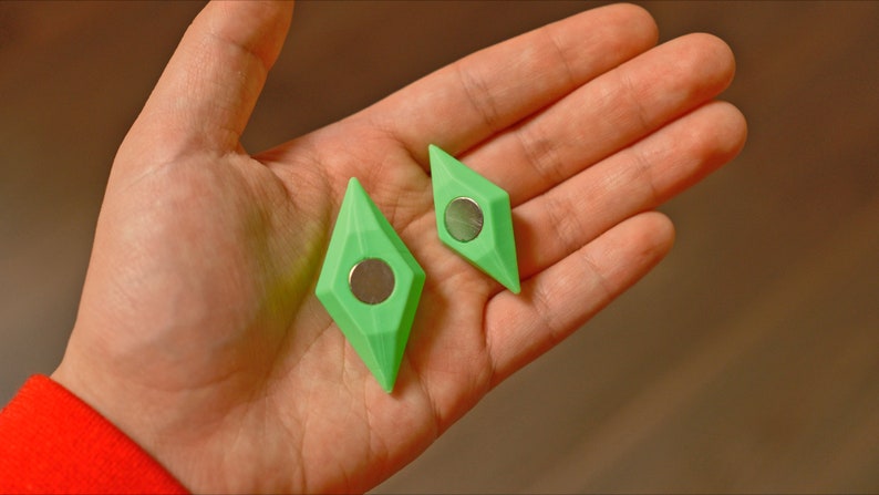 May include: Two green diamond-shaped magnets with a silver center. The magnets are in the palm of a hand.