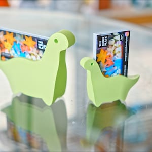 May include: Two green dinosaur-shaped card holders, one larger than the other, with business cards inserted. The cards feature a colourful design and the text "The Build Plate".