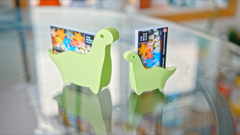 May include: Two green dinosaur-shaped card holders, one larger than the other, with business cards inserted. The cards feature a colourful design and the text "The Build Plate".