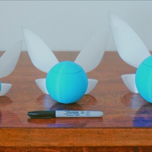 Puede incluir: Tres figuritas de hadas azules y blancas de diferentes tama&ntilde;os. Las figuritas tienen forma de esfera con alas. Las alas son blancas y las esferas son azules. Las figuritas est&aacute;n dispuestas en fila sobre una superficie de madera.
