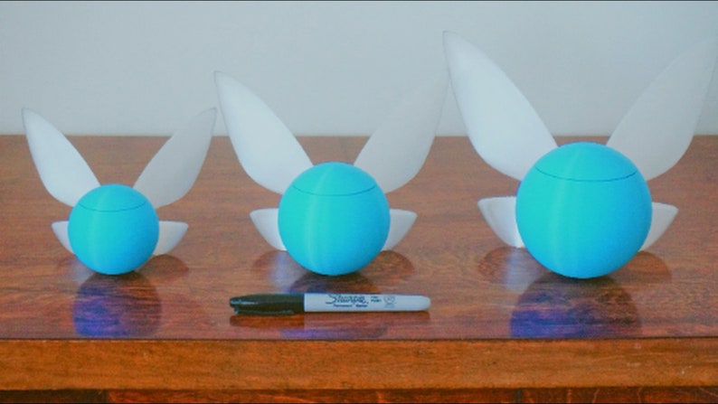 Puede incluir: Tres figuritas de hadas azules y blancas de diferentes tama&ntilde;os. Las figuritas tienen forma de esfera con alas. Las alas son blancas y las esferas son azules. Las figuritas est&aacute;n dispuestas en fila sobre una superficie de madera.