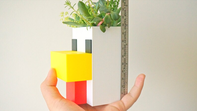 May include: A white, yellow, and red ceramic planter shaped like a pixelated chicken. The planter has a small opening at the top for planting succulents or other small plants. The planter is approximately 4 inches tall.