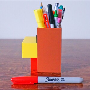 K&ouml;nnte beinhalten: Ein Stiftehalter im Lego-Stil in Braun, Gelb und Rot mit einer Vielzahl von Stiften und Bleistiften darin. Ein roter Sharpie Permanentmarker mit feiner Spitze liegt auf einem Holztisch vor dem Halter.