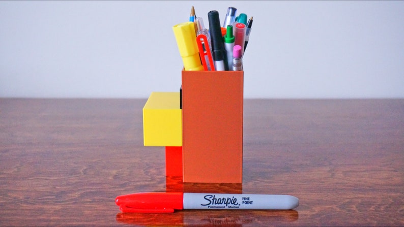 K&ouml;nnte beinhalten: Ein Stiftehalter im Lego-Stil in Braun, Gelb und Rot mit einer Vielzahl von Stiften und Bleistiften darin. Ein roter Sharpie Permanentmarker mit feiner Spitze liegt auf einem Holztisch vor dem Halter.