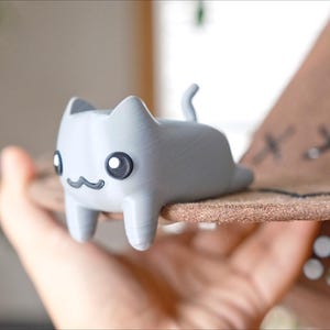 May include: A light grey cat figurine with large black eyes and a small, curved mouth. The cat is lying on a brown hat with decorative stitching. The hat is held in a person's hand.