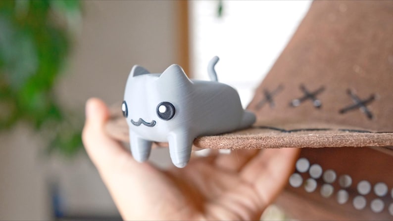 May include: A light grey cat figurine with large black eyes and a small, curved mouth. The cat is lying on a brown hat with decorative stitching. The hat is held in a person's hand.