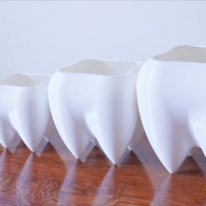 Puede incluir: Un conjunto de cinco macetas de cer&aacute;mica blanca con forma de dientes. Las macetas est&aacute;n dispuestas en fila, disminuyendo de tama&ntilde;o de derecha a izquierda. El acabado liso y brillante refleja la luz, creando una est&eacute;tica limpia y moderna.