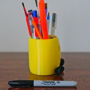 May include: A yellow 3D printed pencil holder shaped like a cartoon character with black feet. The holder is filled with various pens, pencils, and markers, including a red, orange, and blue pen, a black marker, and a silver pen.