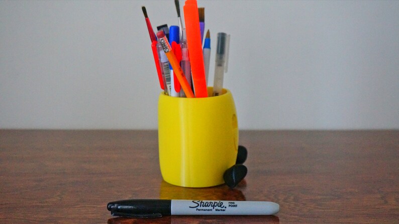 May include: A yellow 3D printed pencil holder shaped like a cartoon character with black feet. The holder is filled with various pens, pencils, and markers, including a red, orange, and blue pen, a black marker, and a silver pen.