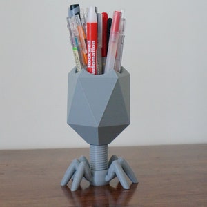 May include: A gray, geometric pen holder shaped like a bacteriophage with six legs. The holder is filled with a variety of pens, including red, black, and blue.