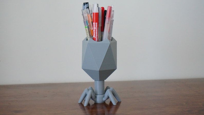 May include: A gray, geometric pen holder shaped like a bacteriophage with six legs. The holder is filled with a variety of pens, including red, black, and blue.